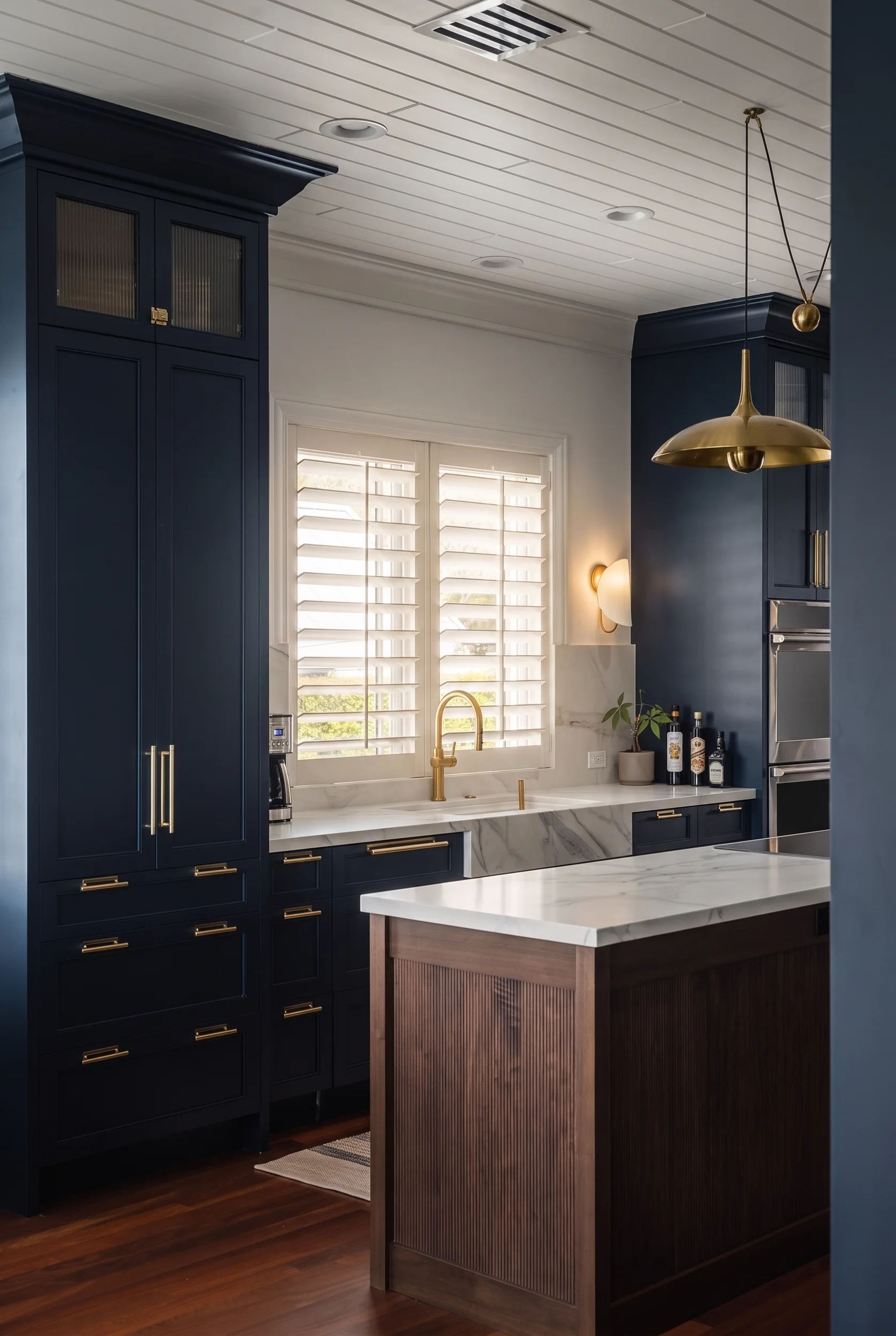 Vertical detail of a navy kitchen with a brass pendant, plantation shutters, and a marble-topped island