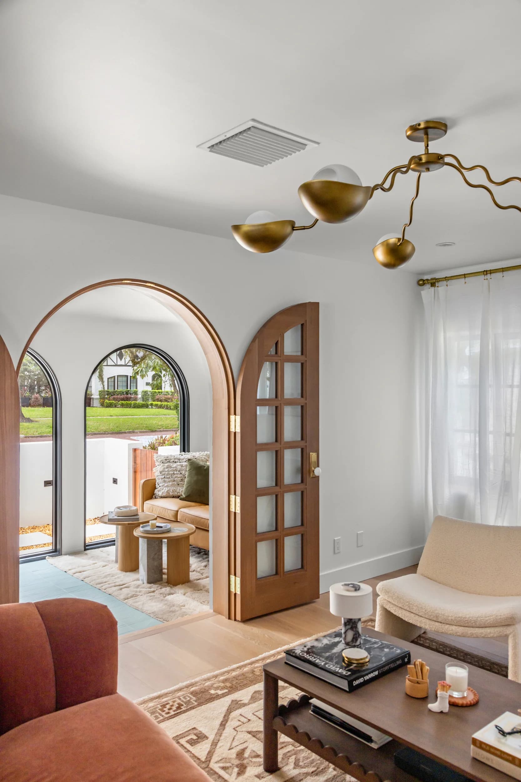 View through arched wood French doors into a sunlit living room with a rust velvet sofa