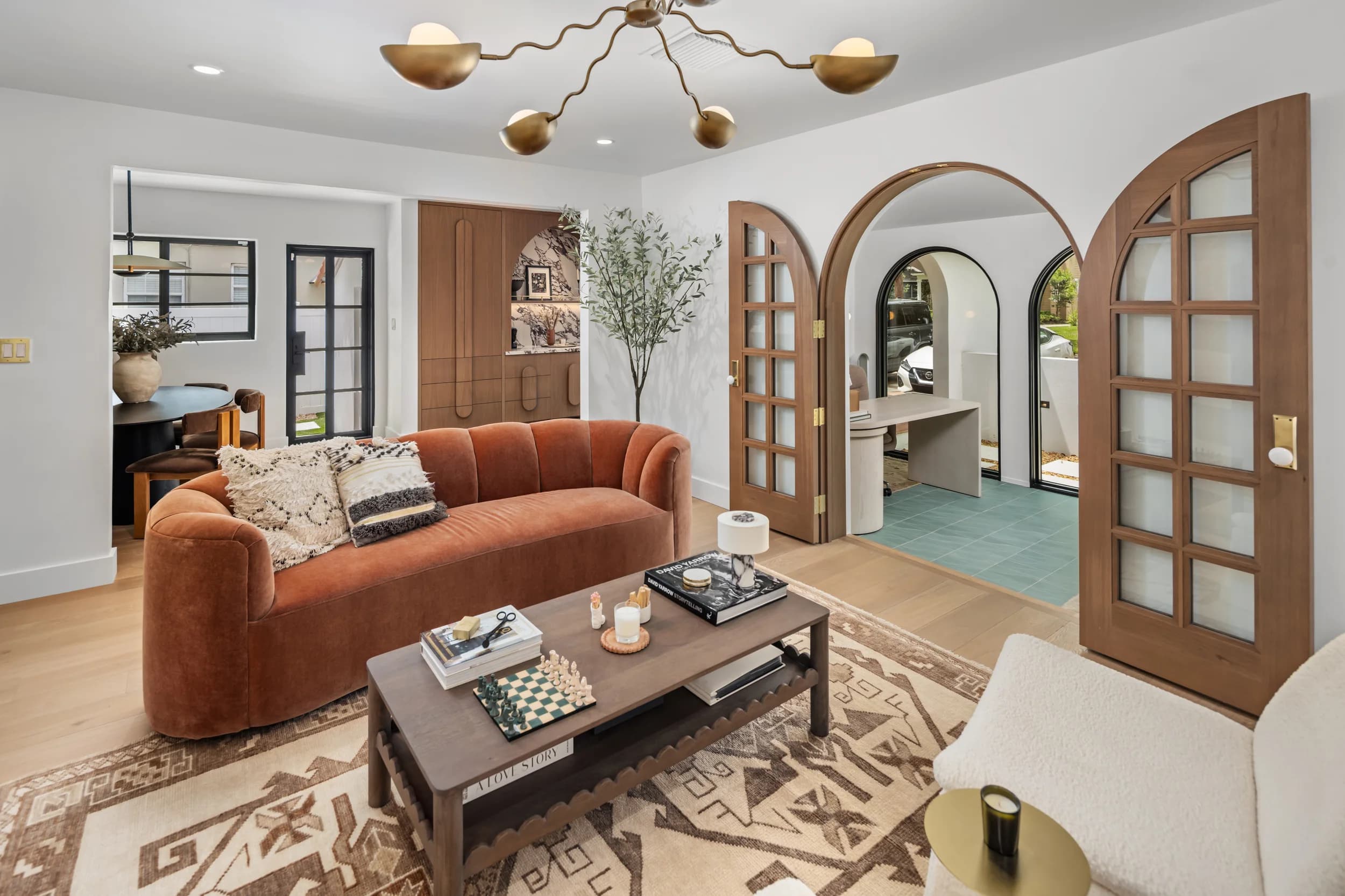 Living room styled with a rust velvet sofa, carved wood coffee table, boucle chair, and brass chandelier beneath arched doorways