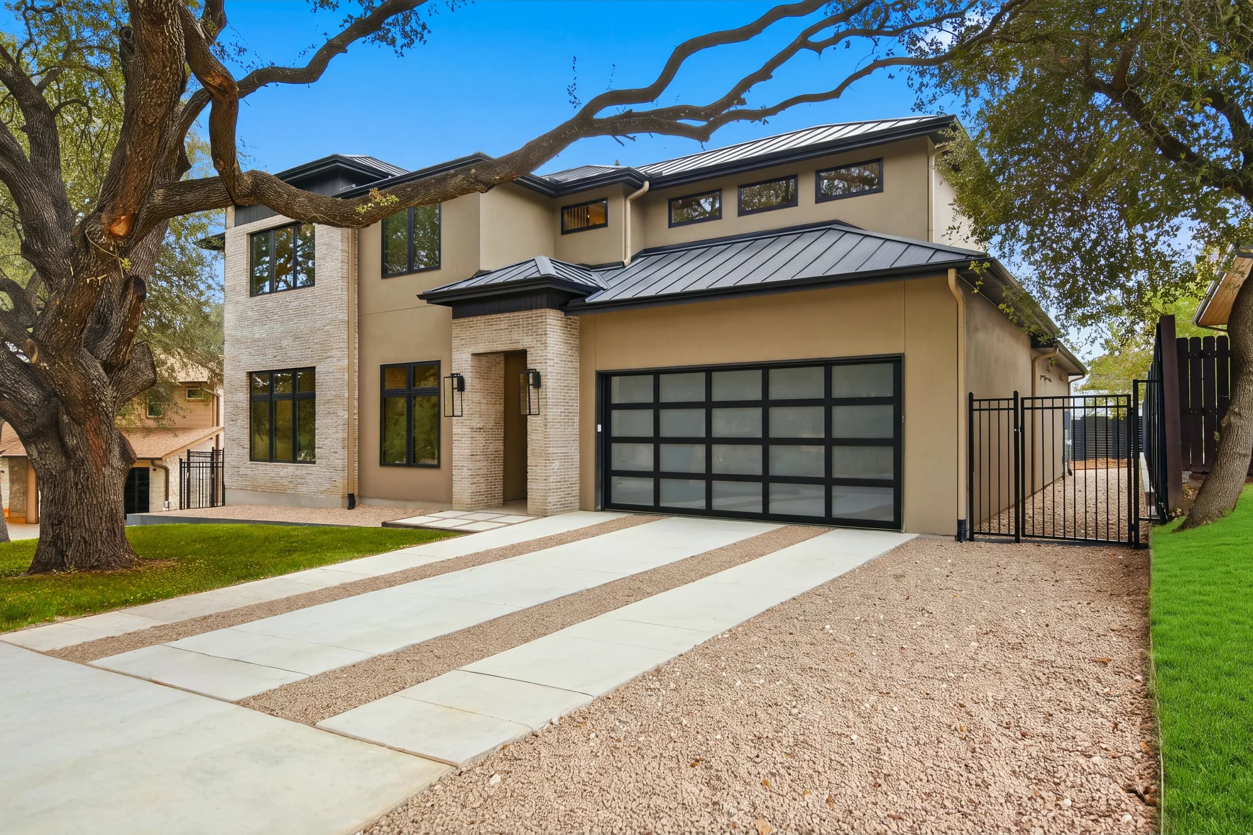 Modern two-story home with brick and stucco, standing-seam metal roof and glass garage door under a live oak