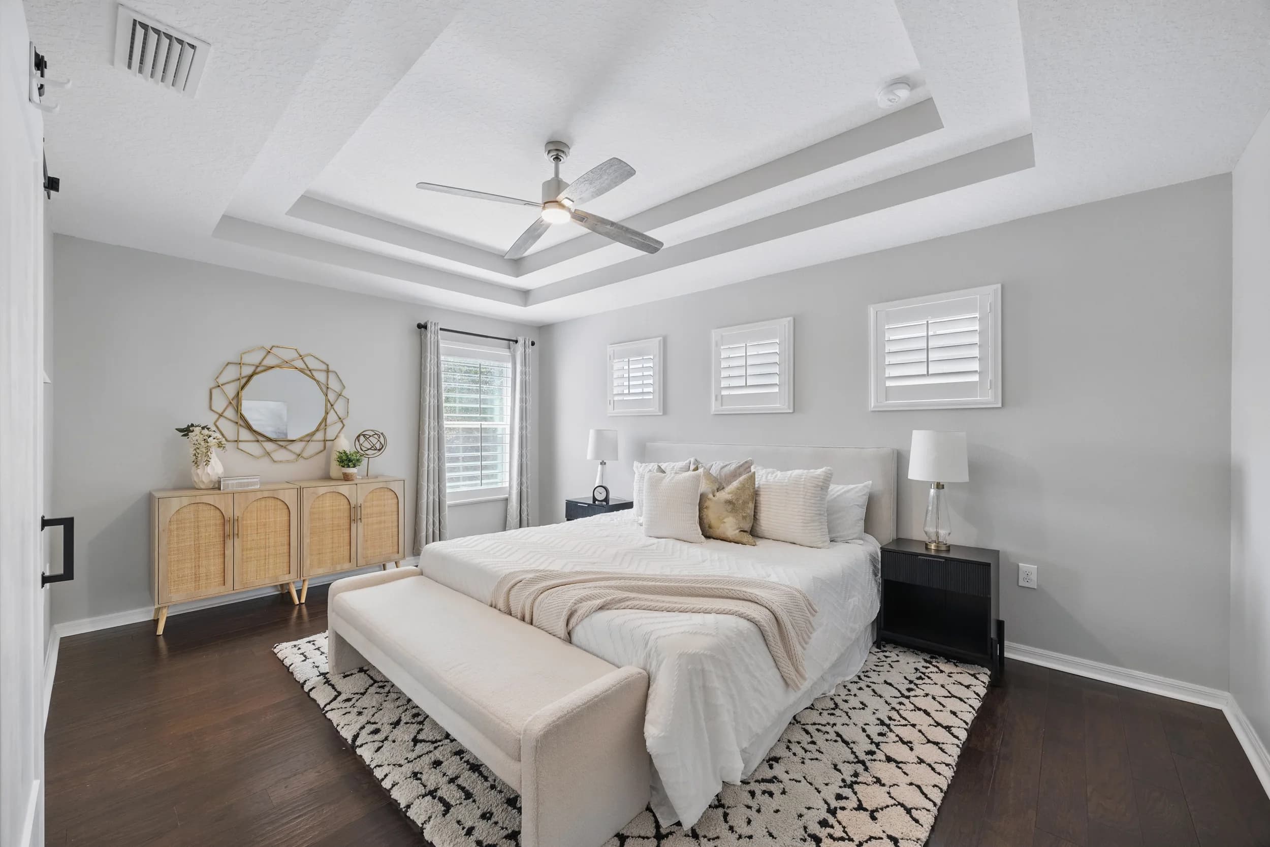 Primary bedroom with tray ceiling, rattan dresser, sunburst mirror and patterned rug