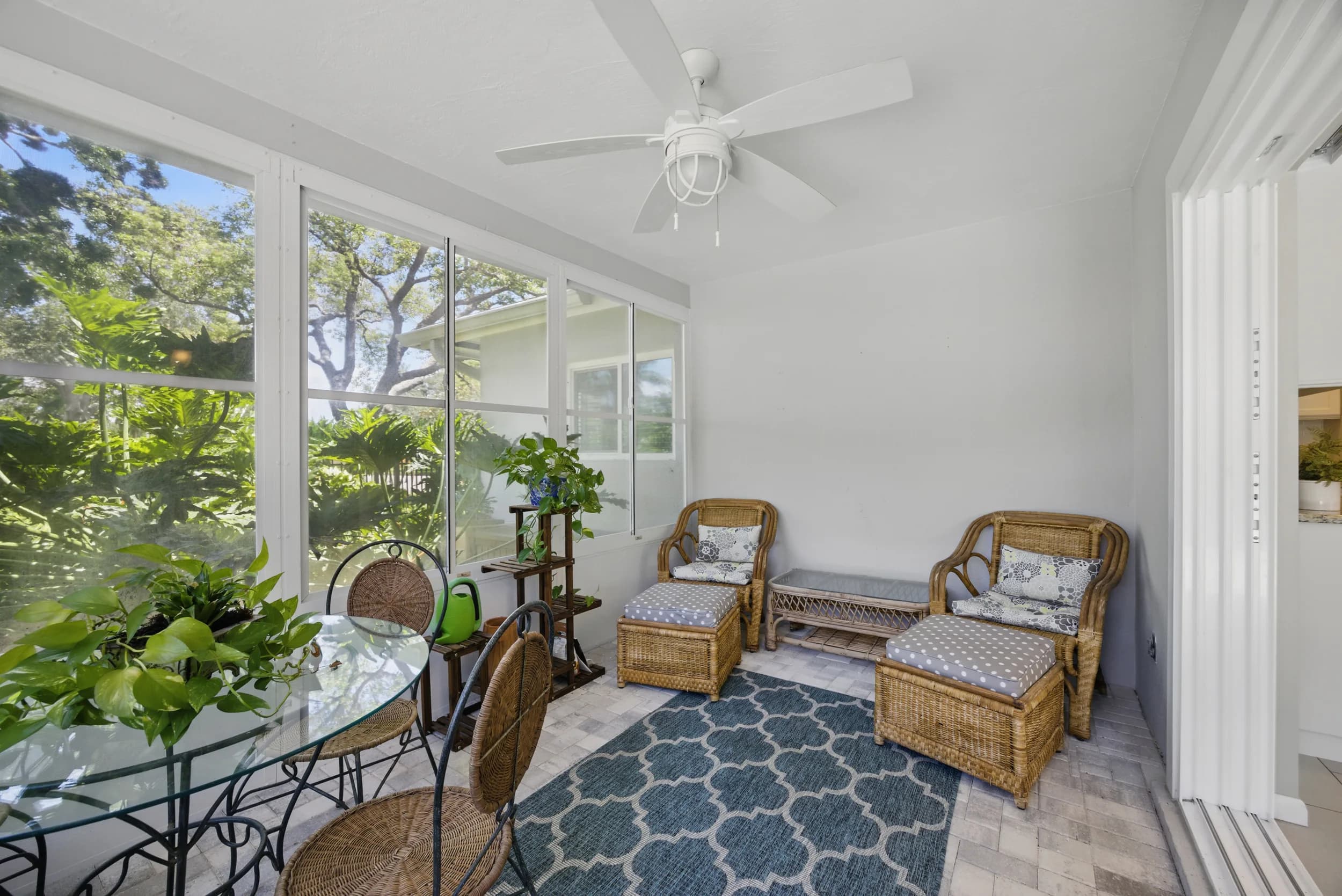 Sunroom staged with wicker chairs, ottomans and a glass bistro table surrounded by greenery