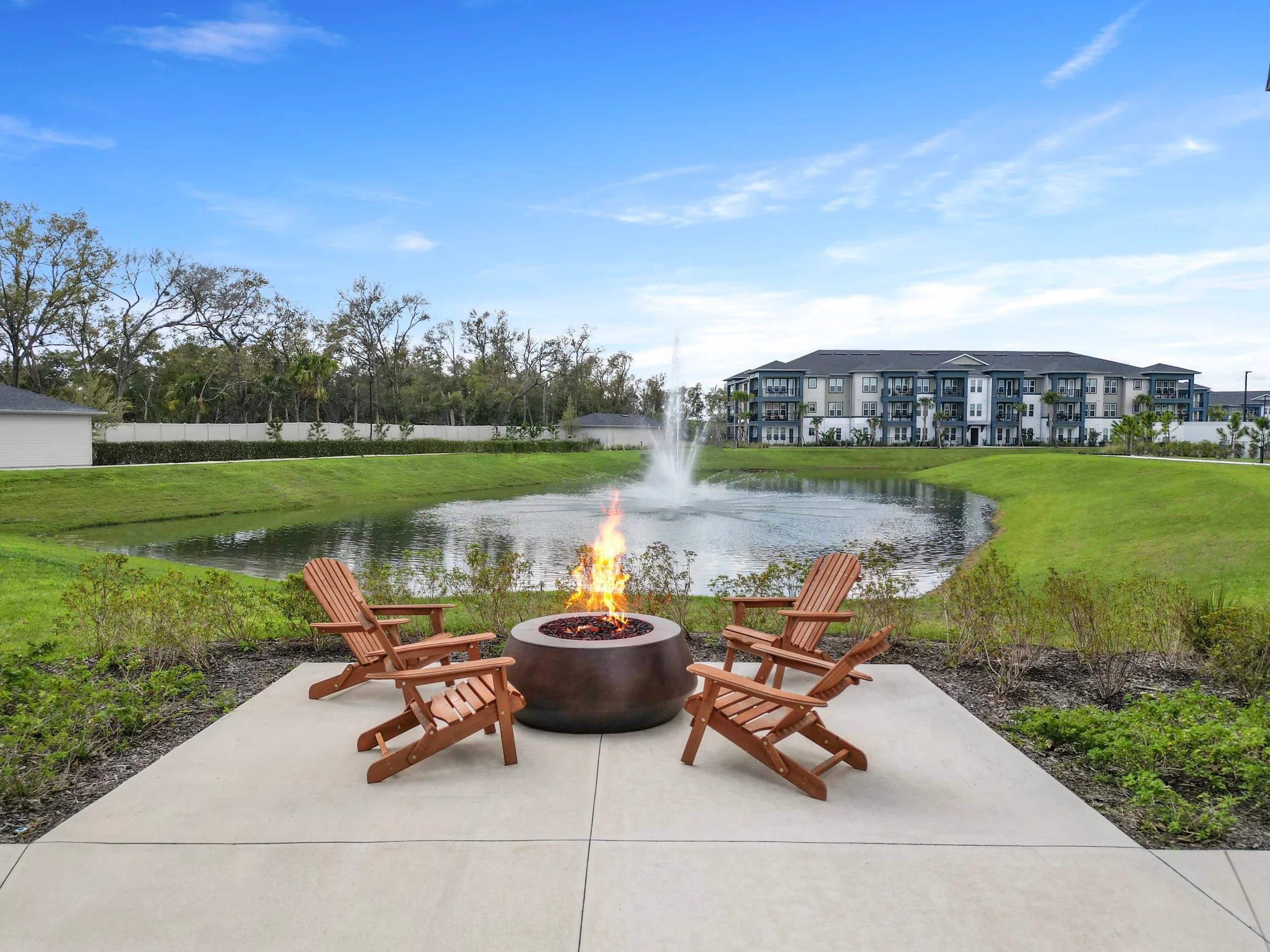 Fire pit with Adirondack chairs beside a community lake and fountain