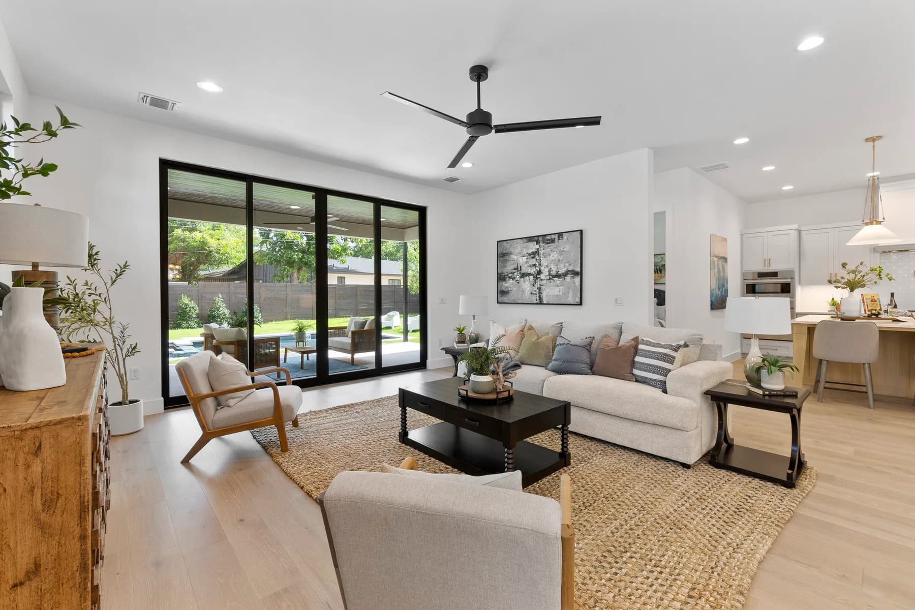 Modern living room with a beige sofa, two chairs, and a black coffee table, leading to a kitchen area. Large sliding glass doors open to an outdoor pool. The space features bright lighting and minimalistic decor.