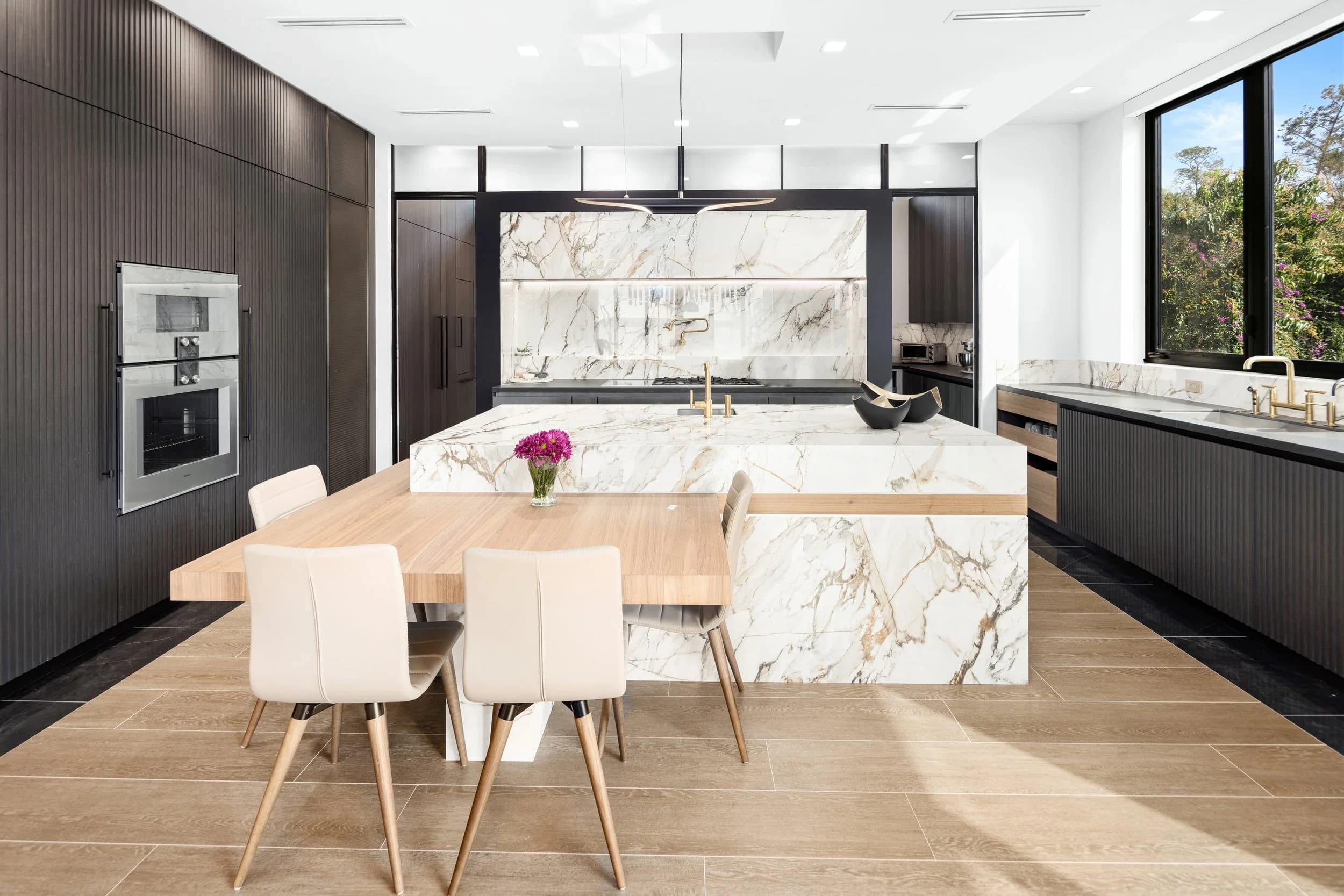 Kitchen with book-matched marble waterfall island, attached wood dining table and dark cabinetry
