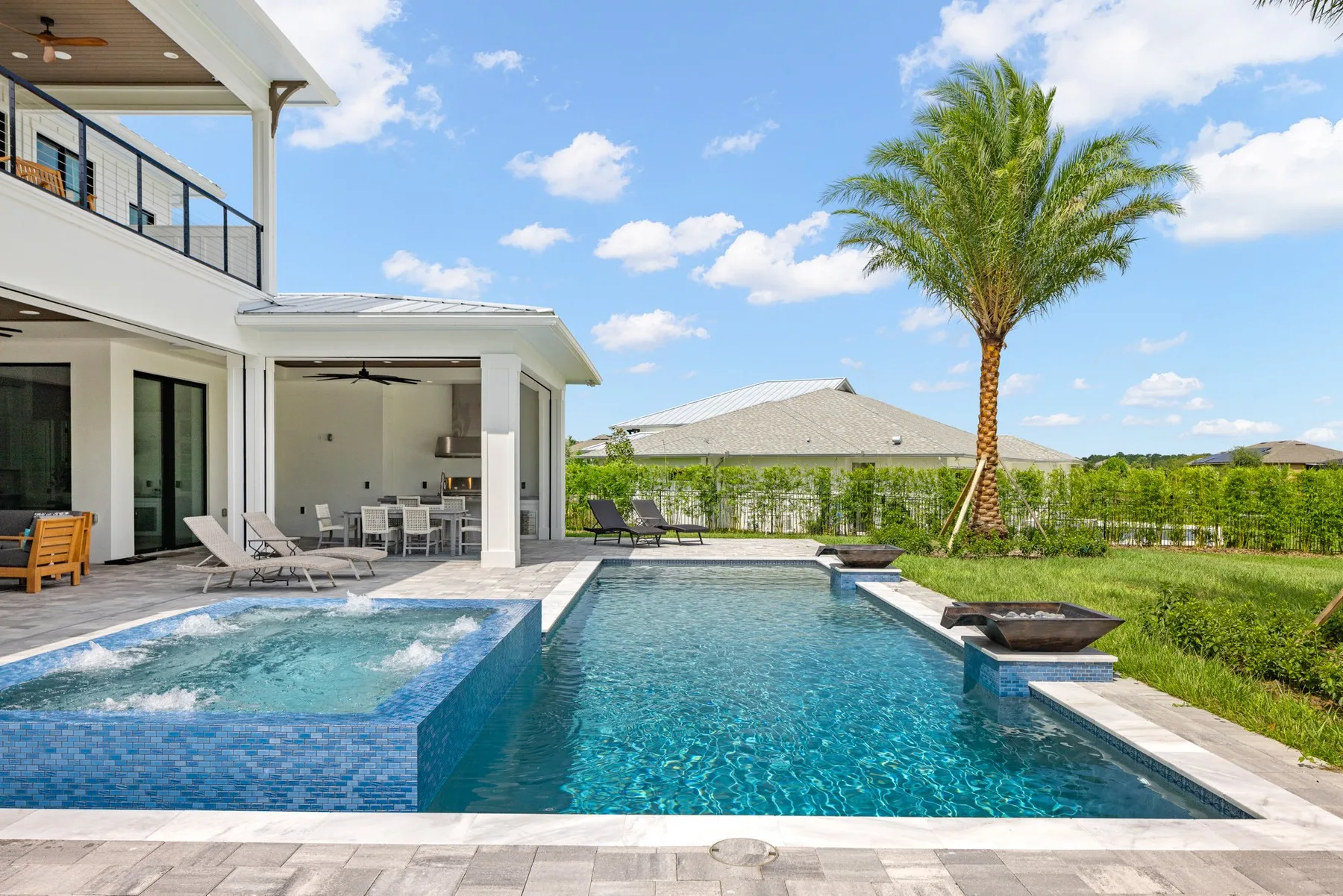 Exterior view of a luxurious residential backyard with a swimming pool, spa and an outdoor kitchen under a covered patio. The area is bordered by a neatly maintained lawn and a single palm tree, with a clear sky above.