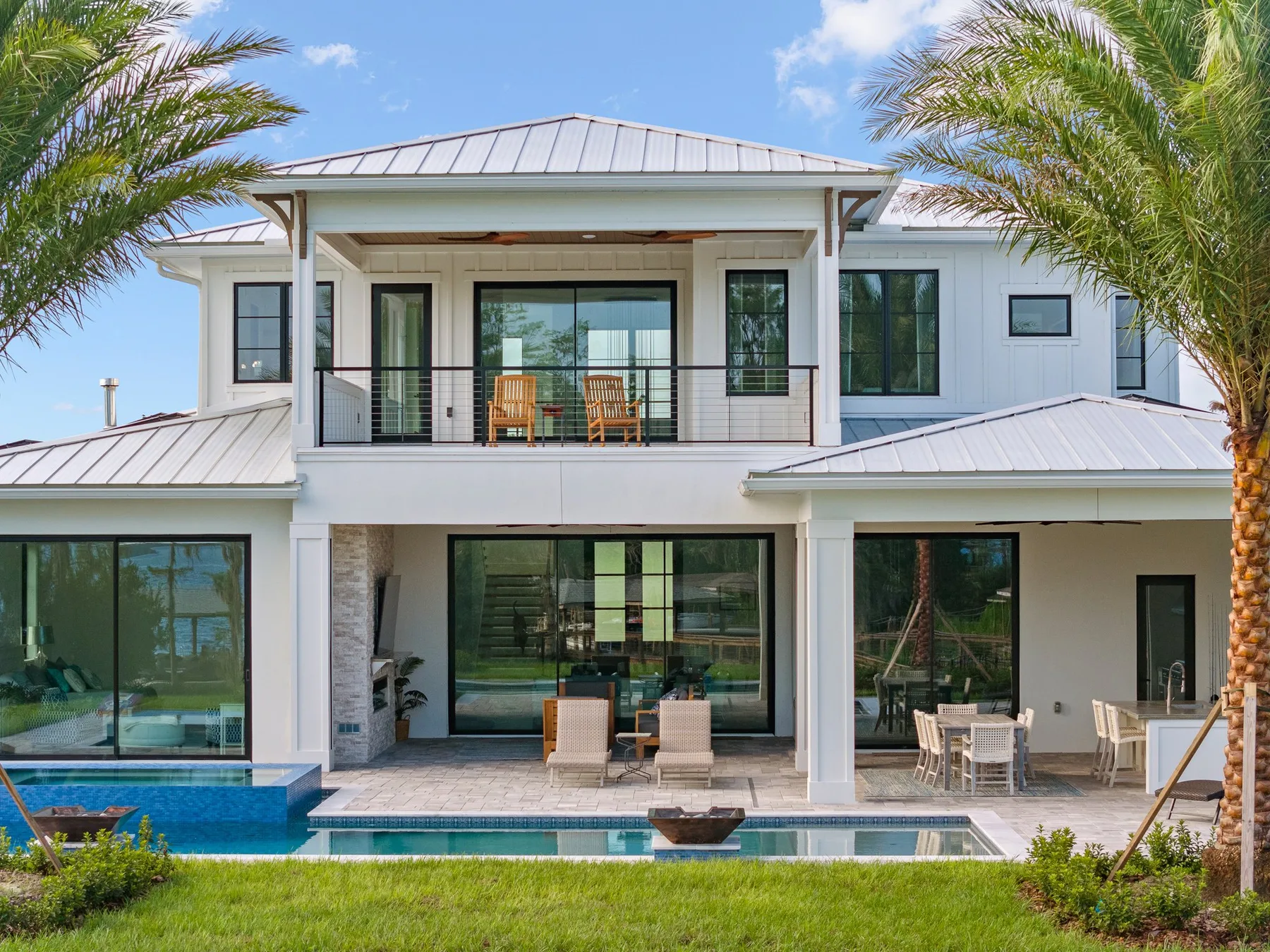 Luxurious two-story house with expansive glass windows, a balcony, swimming pool, and surrounding palm trees.