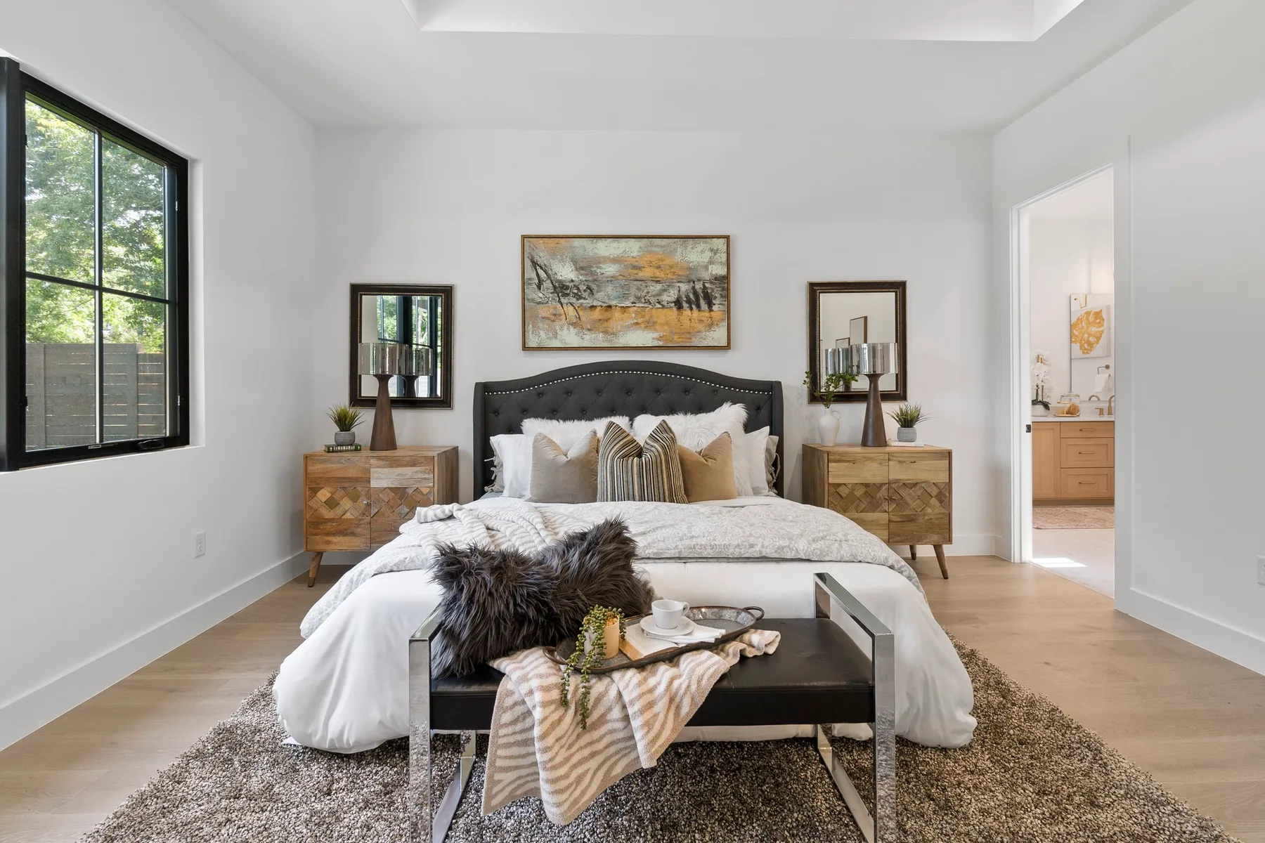 Modern bedroom featuring a large bed with a tufted headboard and a variety of textured pillows. The room includes two nightstands, a bench at the foot of the bed, and an abstract painting above the headboard. Large windows provide ample natural light, and the space is decorated in neutral tones.