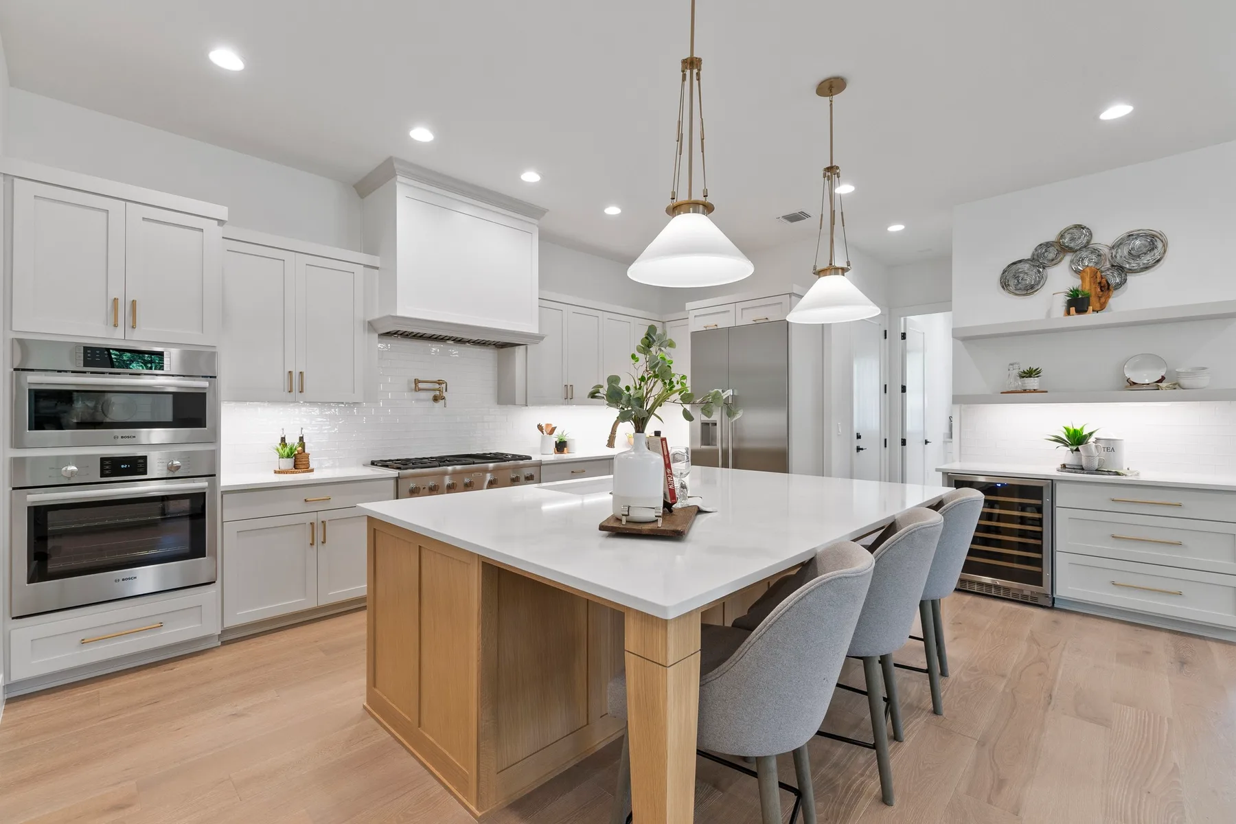Modern kitchen with white cabinetry and a central island featuring light wood accents. Includes built-in appliances and hanging pendant lights. Decorative elements include a floral centerpiece and wall art.