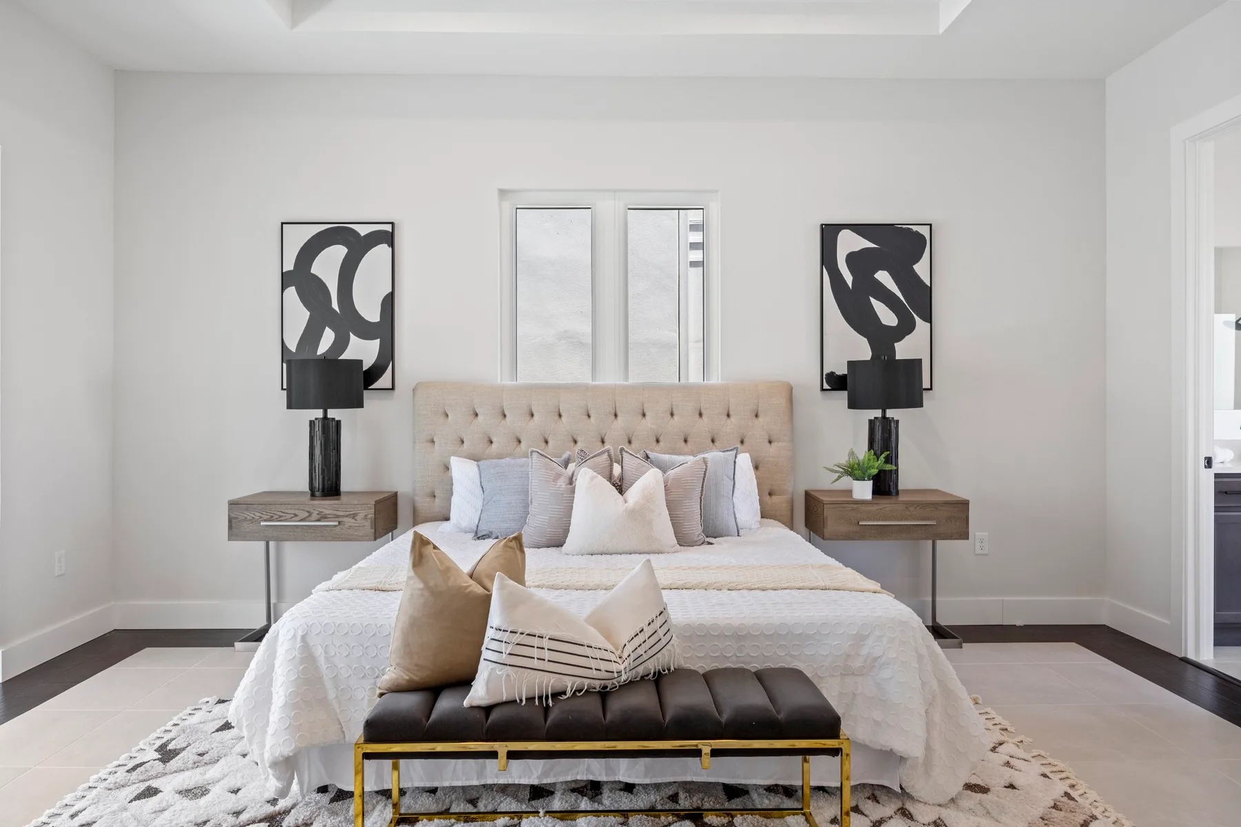 Modern bedroom featuring a large tufted headboard with a bed adorned with neutral-toned bedding and multiple pillows. Two symmetrical nightstands with abstract black sculptures and black table lamps flank the bed. A black bench sits at the foot of the bed, and the room has light gray walls and white trimming.