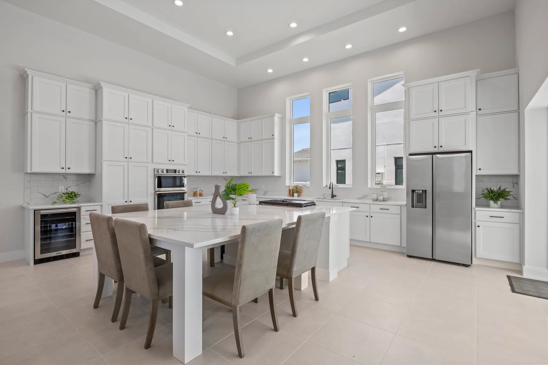 Modern kitchen interior featuring white cabinetry, stainless steel appliances (including a refrigerator and oven), a central island, and a dining table with chairs. Natural light streams in through large windows.